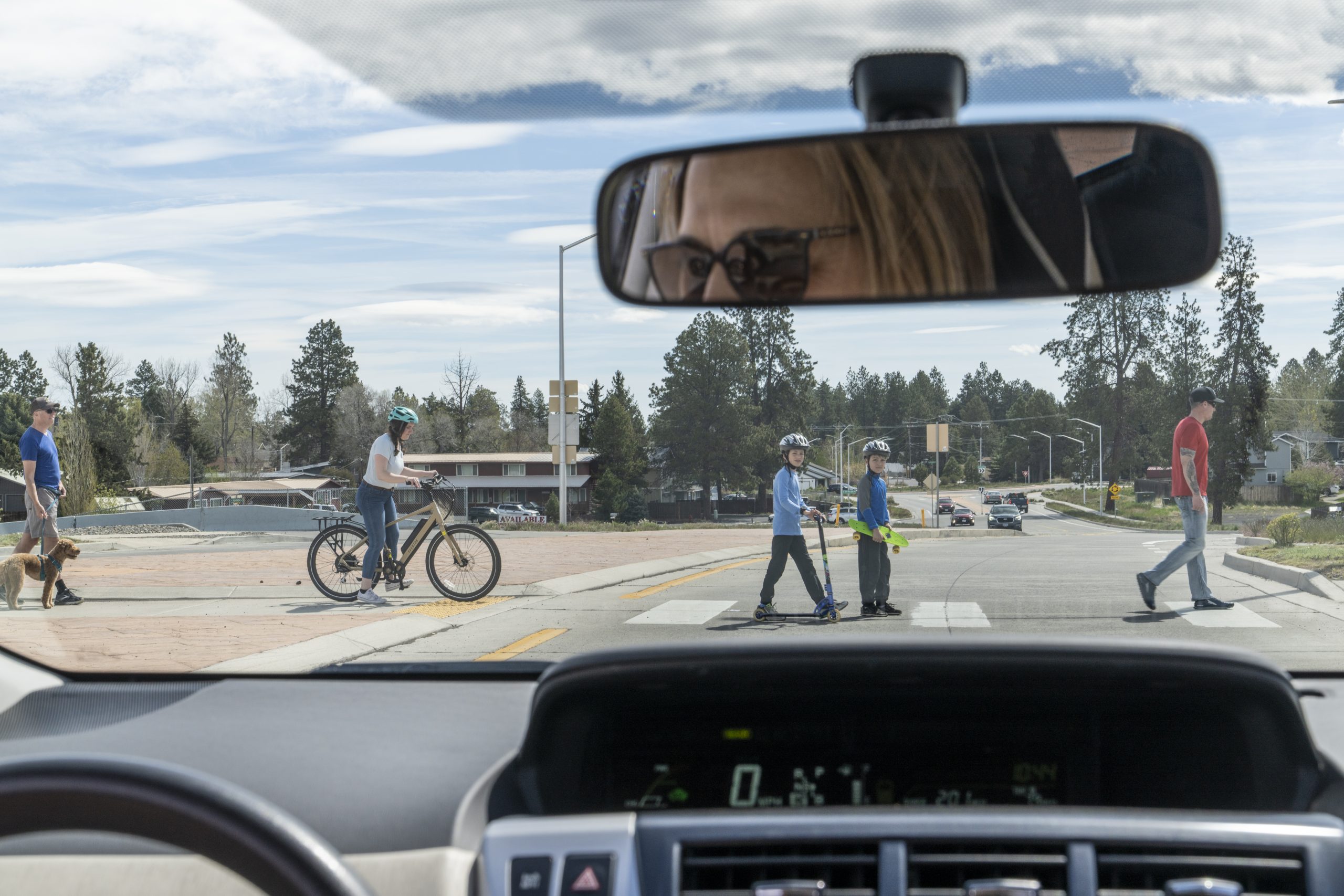 Image showing the view from inside a car, with a driver looking through the rearview mirror at pedestrians and cyclists crossing the street, emphasizing the importance of driver education in fostering awareness and safe interactions with all road users.