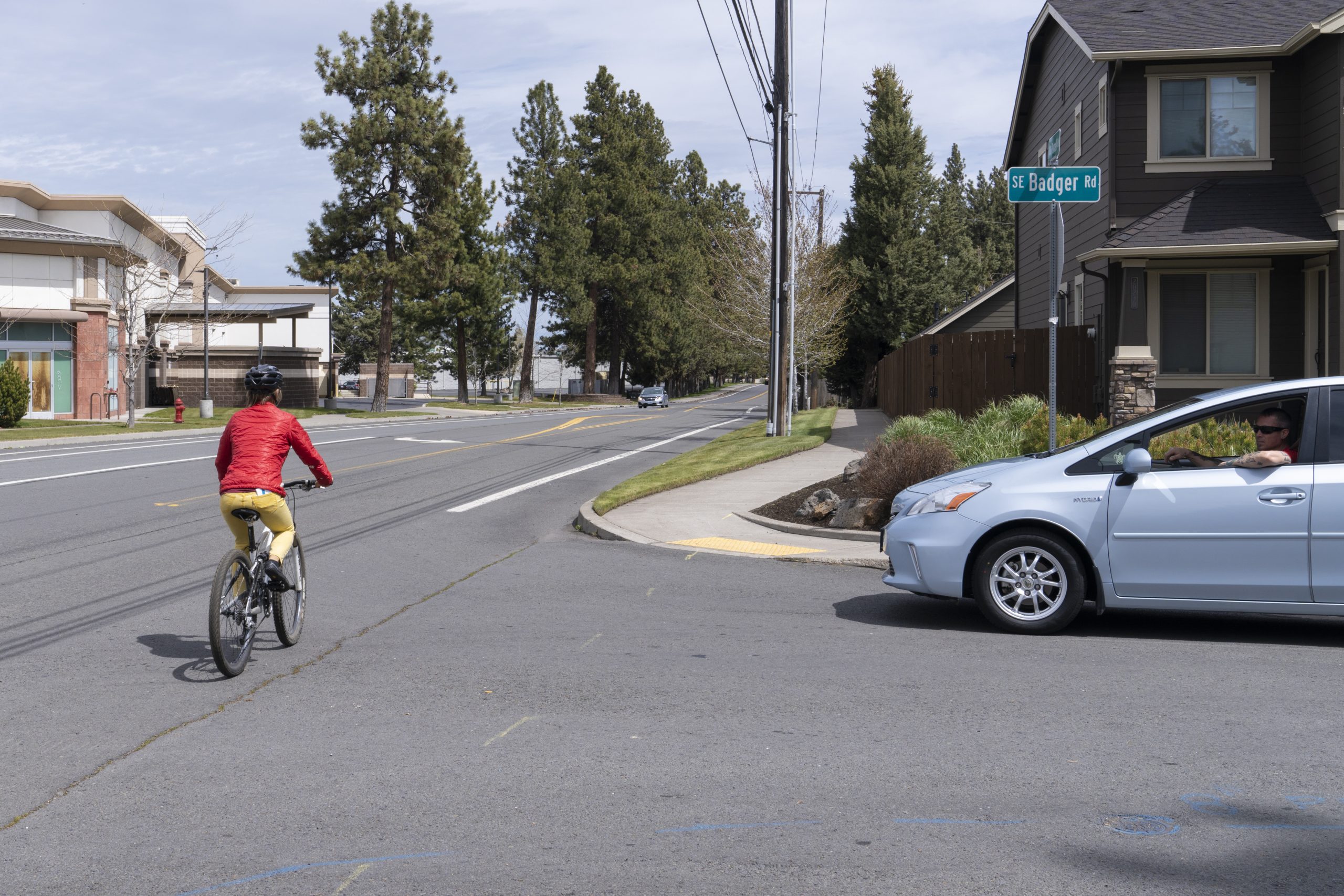 A cyclist in a red jacket rides past a silver car waiting at a street intersection.