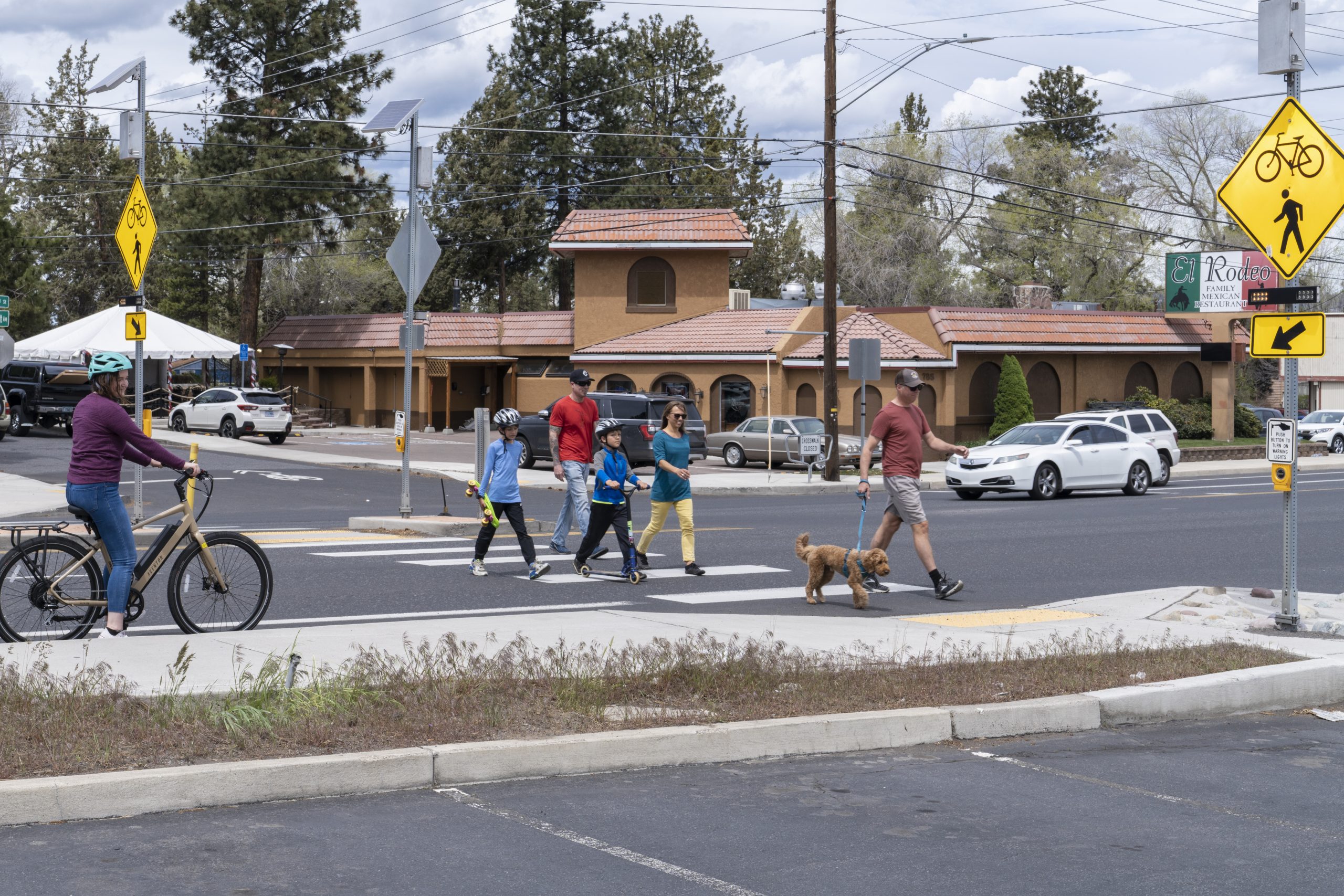 A group of pedestrians, including a cyclist and a person with a dog, cross a street near traffic signs in Bend, OR. The scene is related to free driving classes.