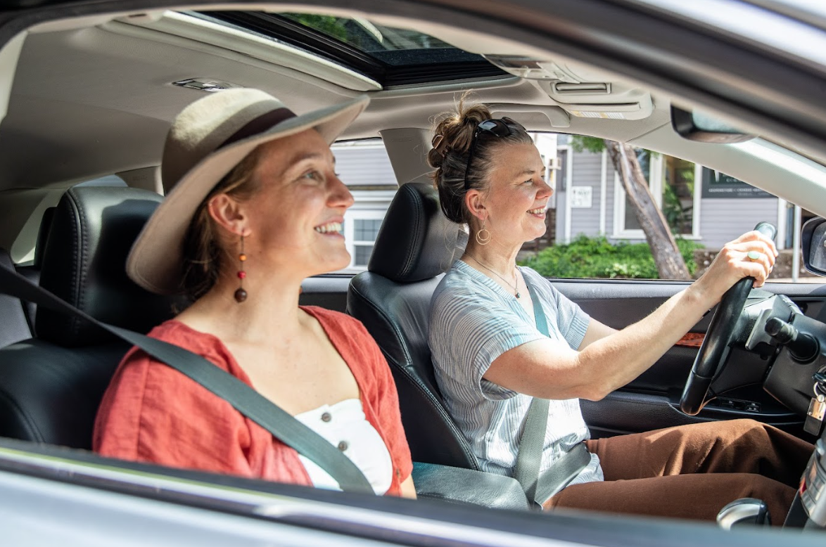 Image of two women in a car, one driving and the other sitting in the passenger seat, both smiling, highlighting the importance of using driver safety resources to ensure safe and confident driving for all road users.