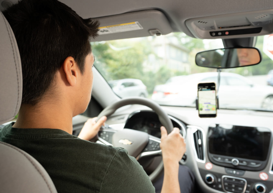Person driving a car, holding the steering wheel with a phone mounted on the dashboard showing a map.