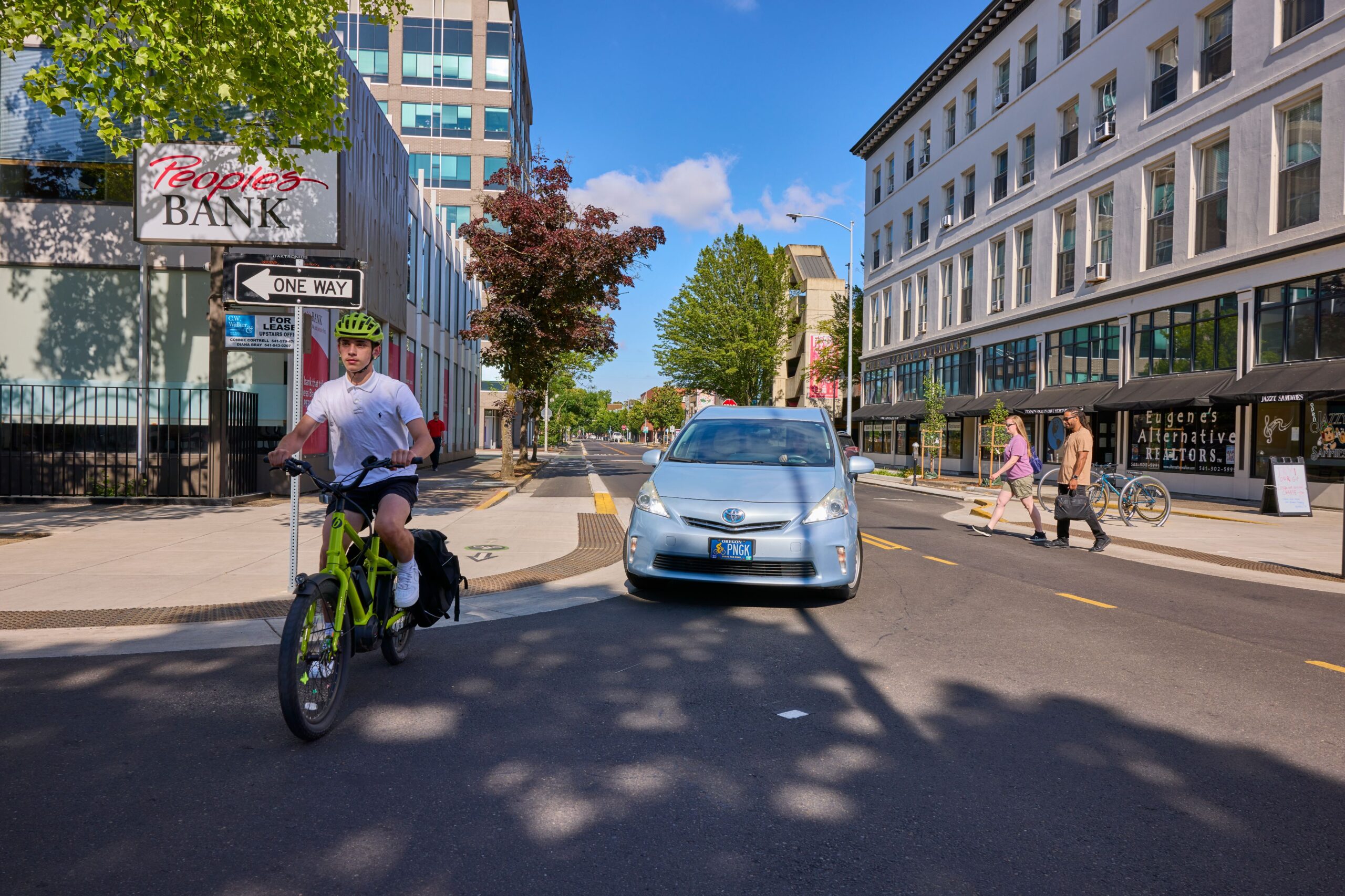 A cyclist riding on a street near a one-way sign, with a car driving past and pedestrians walking in the background. The scene highlights vulnerable road users.