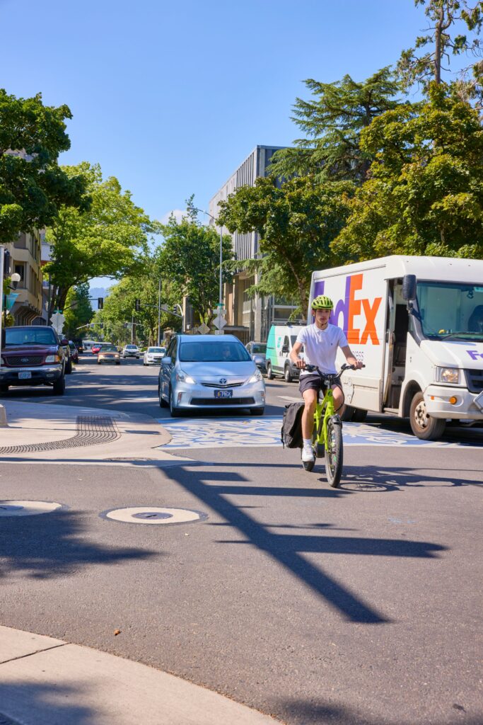 A cyclist riding on a designated bike lane with a FedEx truck and a car passing by in the background.