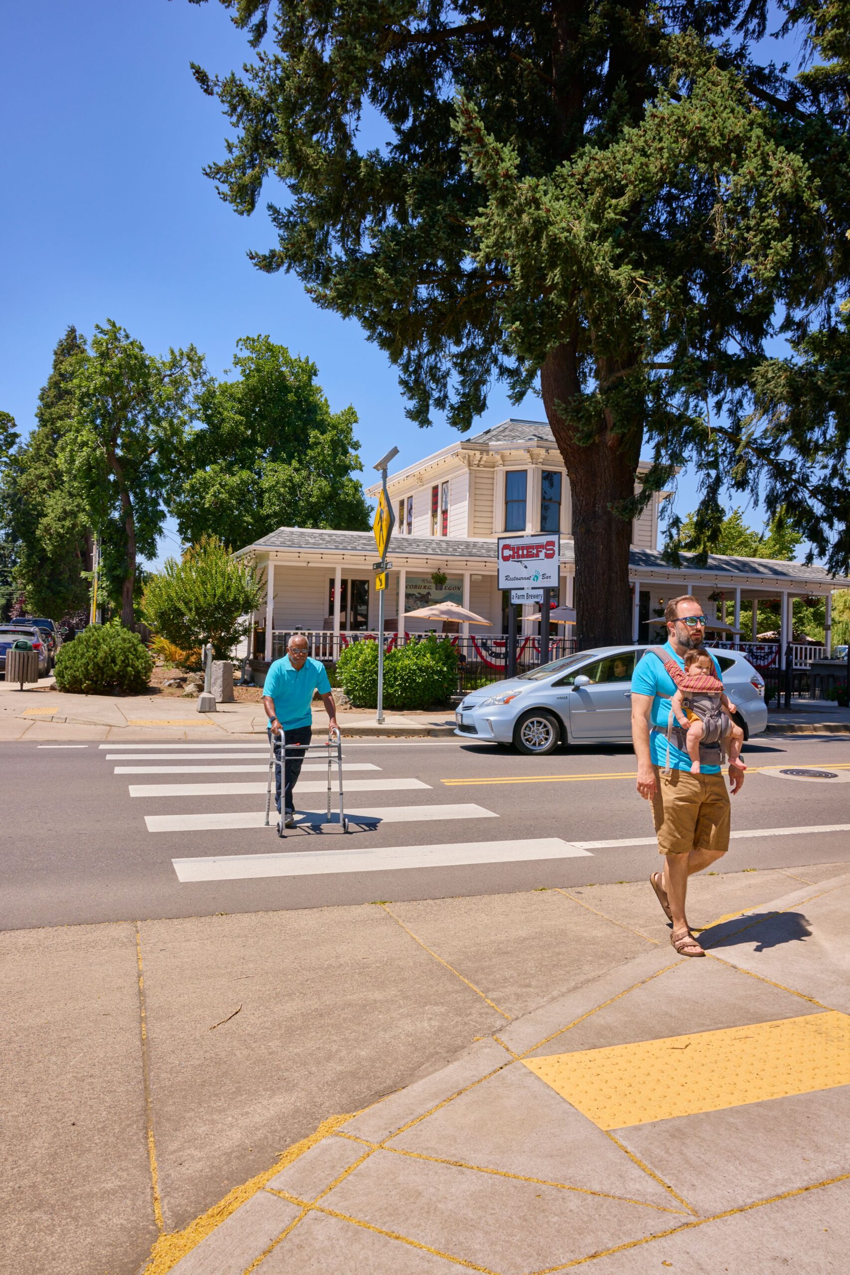 Two pedestrians crossing a street at a crosswalk, with one person using a walker, and a car passing by in the background.