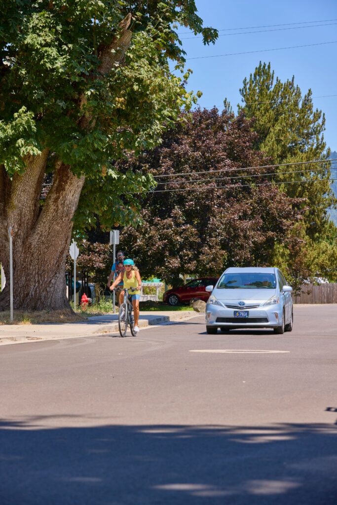 A cyclist riding near a car on the road under trees in Gresham, OR. The scene is related to an Oregon friendly driver course in Gresham, OR.