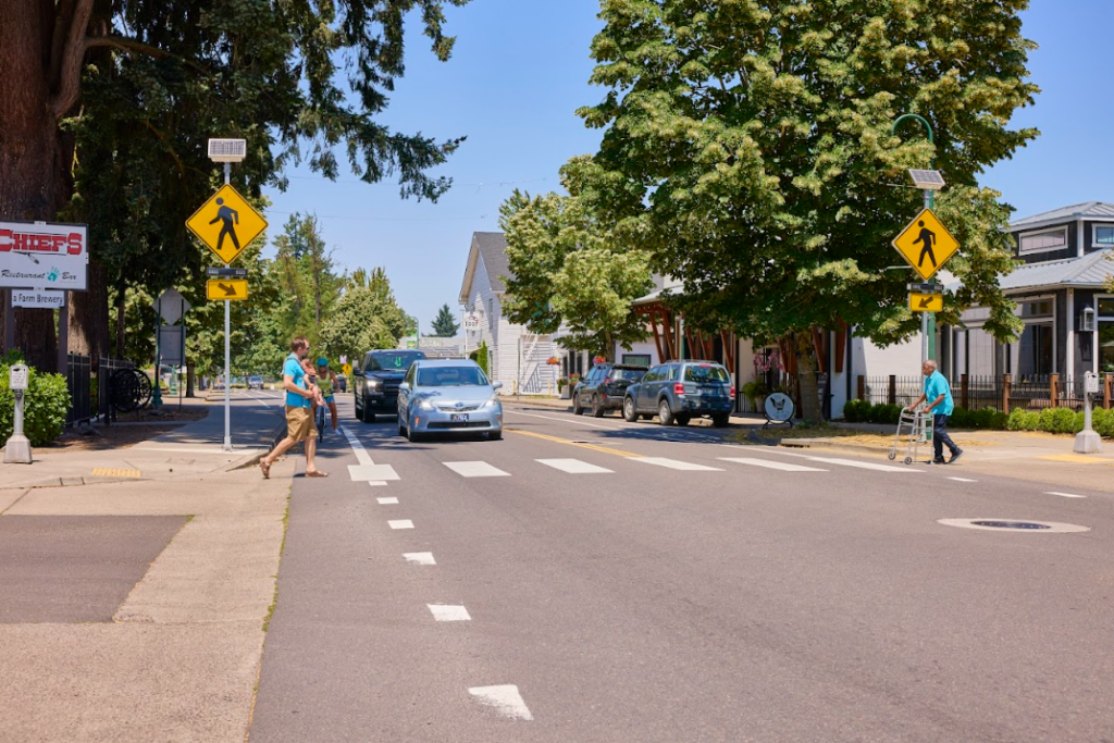 Image showing pedestrians crossing a street with clear pedestrian signs, while a car passes through in Beaverton, OR, emphasizing the importance of a road safety course in Beaverton, OR, for enhancing awareness and safe road practices for both pedestrians and drivers.