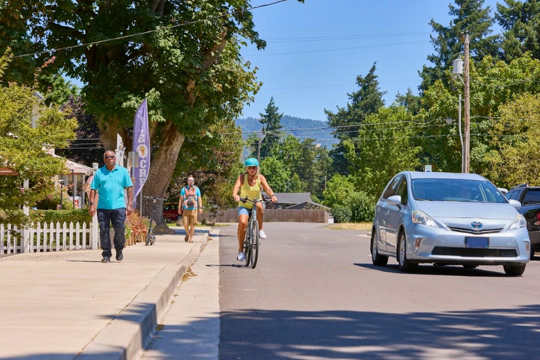 Image depicting pedestrians walking and a cyclist riding along a street, with a car passing by, highlighting the importance of driver safety training for business leaders and teams to ensure safe road practices in everyday environments.