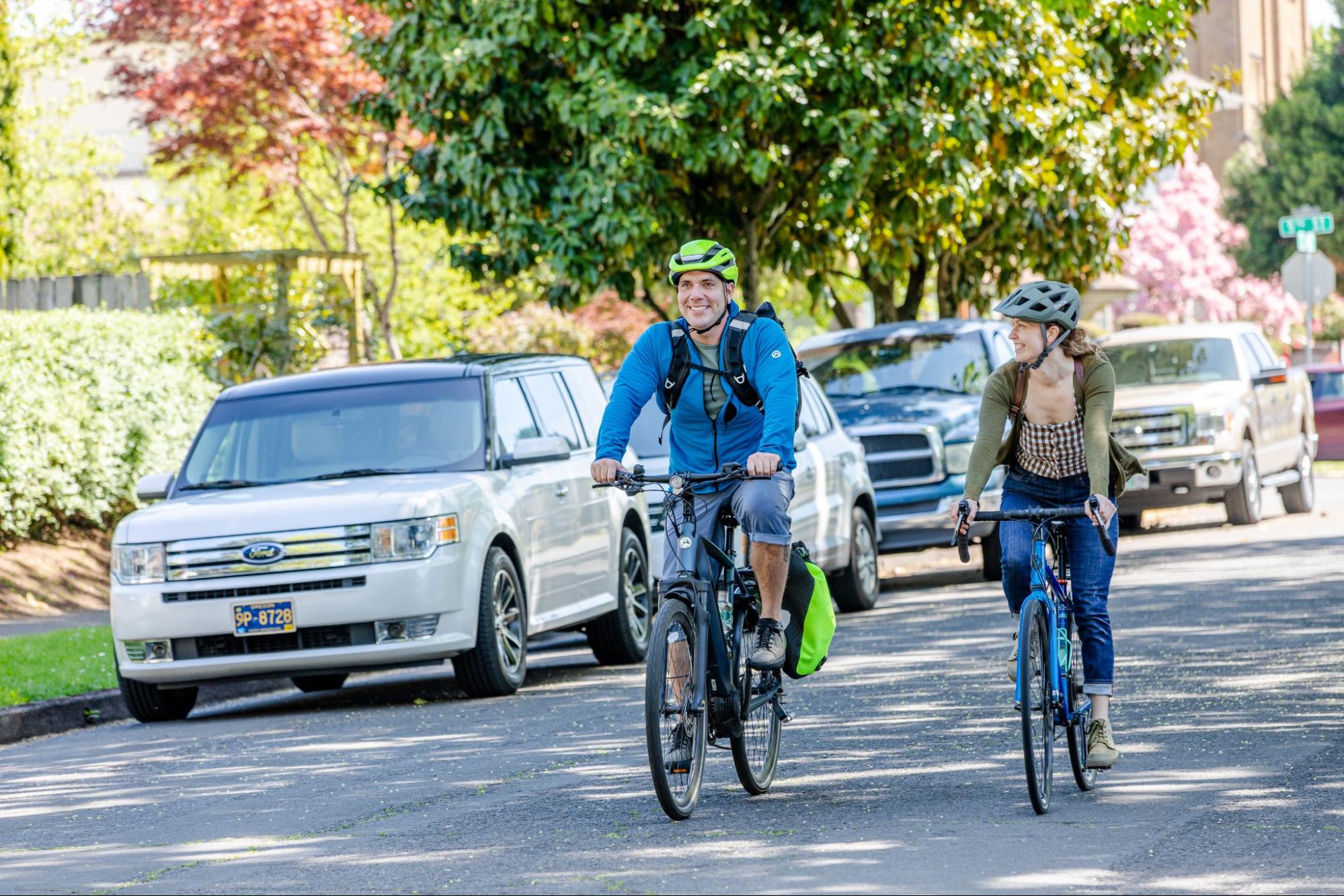 A man and a woman cycle side by side on a street in Springfield, OR. Both are smiling and wearing helmets, the man in a blue shirt and the woman in a green top.