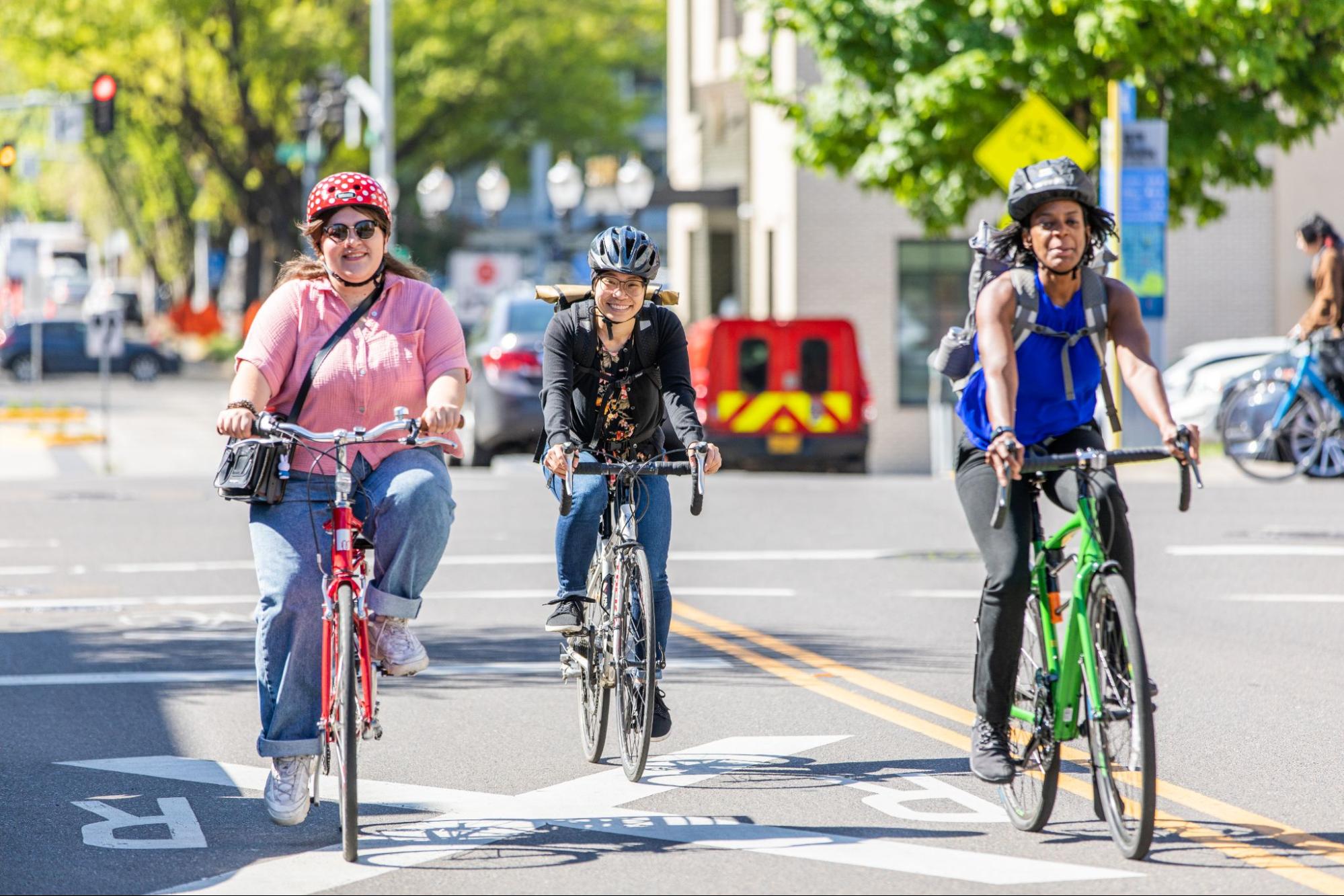 Three cyclists, all wearing helmets, ride together along a bike lane in Springfield, OR. One woman is on a red bike, another on a black bike, and a third woman rides a green bike.