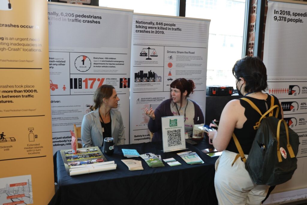 Image showing a booth at an event with two women engaging in conversation with a visitor, surrounded by informative displays and brochures, representing transportation advocates in promoting safe driving and raising awareness about road safety issues.