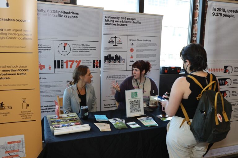 Image showing a booth at an event with two women engaging in conversation with a visitor, surrounded by informative displays and brochures, representing transportation advocates in promoting safe driving and raising awareness about road safety issues.