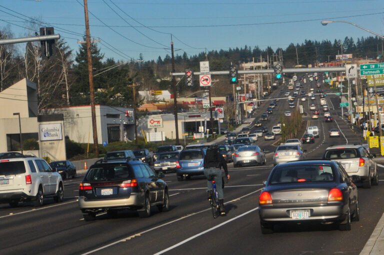 A street scene in Portland showing a cyclist riding along a road with a line of cars, including commercial vehicles. The image illustrates the interaction between commercial drivers and cyclists in urban traffic.