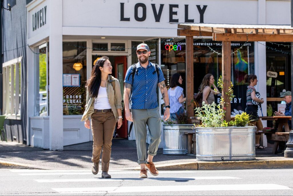 A man and woman walk together across the street in front of a café named "Lovely" in Springfield, OR, with the café's sign visible above them. The woman wears a beige jacket and the man a blue shirt with a backpack.