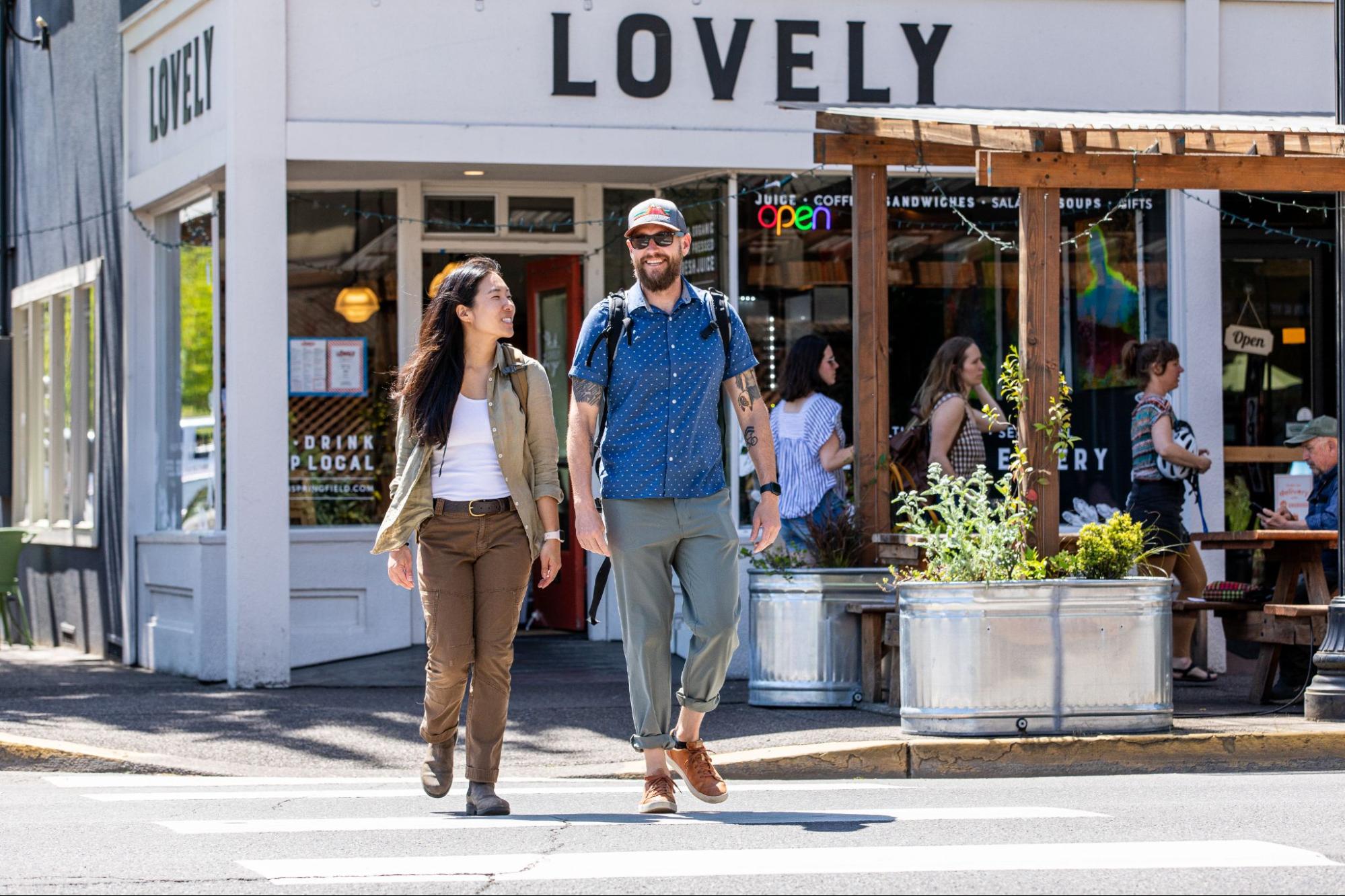 A man and woman walk together across the street in front of a café named "Lovely" in Springfield, OR, with the café's sign visible above them. The woman wears a beige jacket and the man a blue shirt with a backpack.