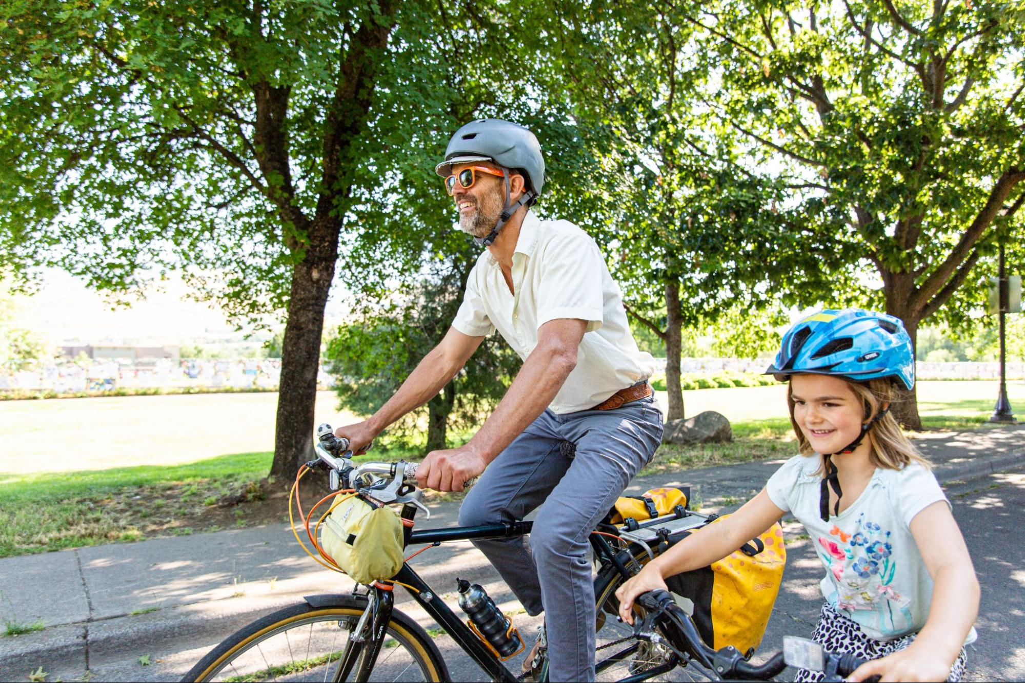Image showing a man and a young girl riding bicycles together in a park, both wearing helmets, highlighting the importance of a road safety course in Medford, OR, to promote safe cycling and awareness of road safety for all users.