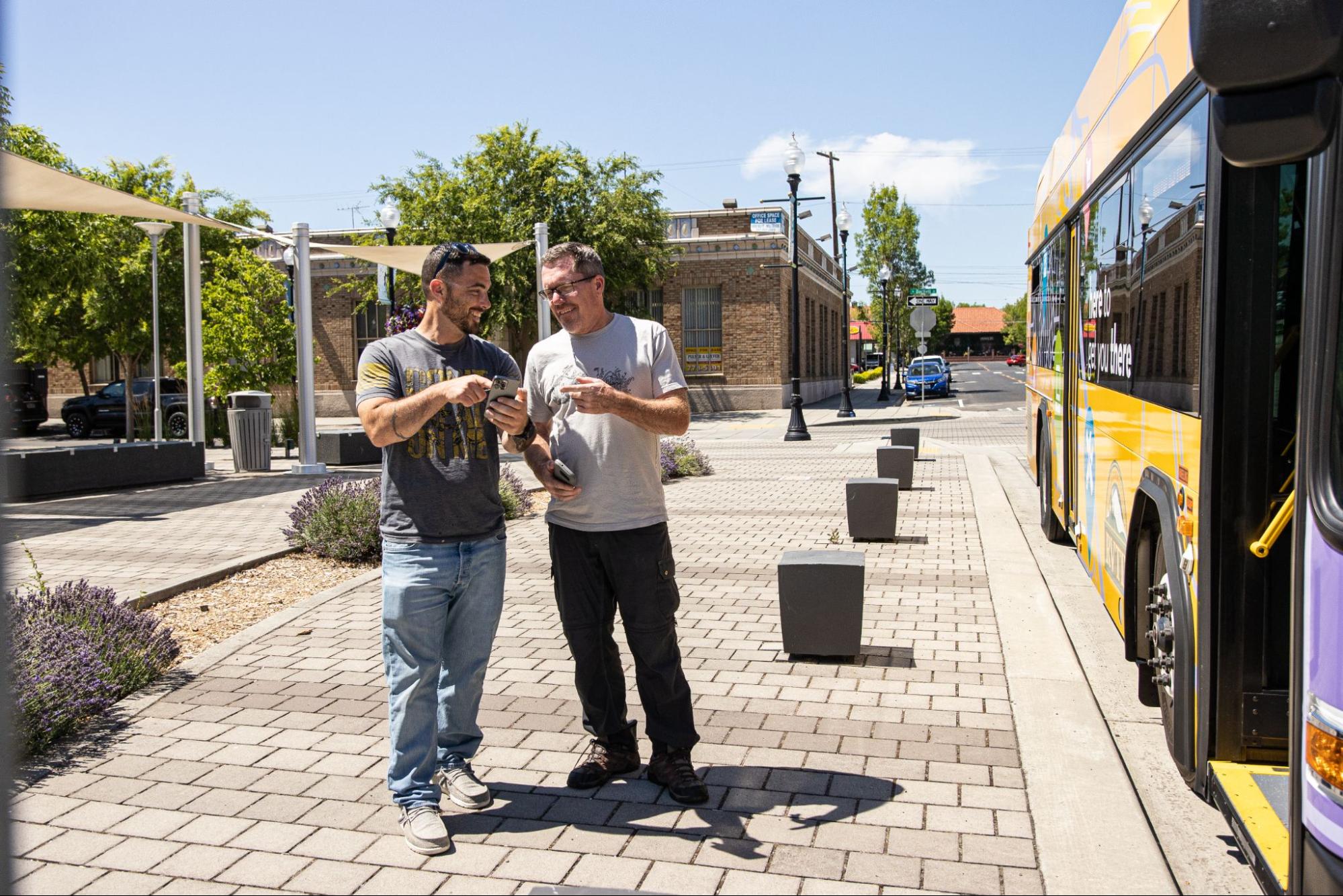 Image showing two men standing near a bus in a sunny urban area, looking at their phones, emphasizing the importance of driving in medford and promoting safe transportation practices for all road users in the community.